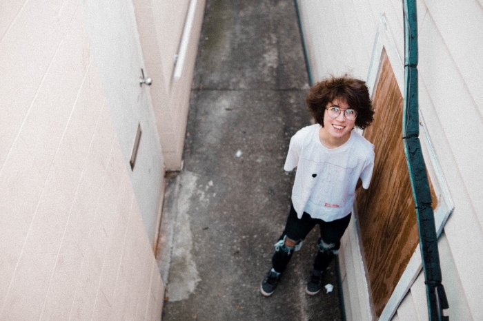 Young adult with short brown hair and eyeglasses wearing a white t-shirt and jeans is smiling at the camera from an overhead viewpoint, while standing in an outdoor alleyway. 