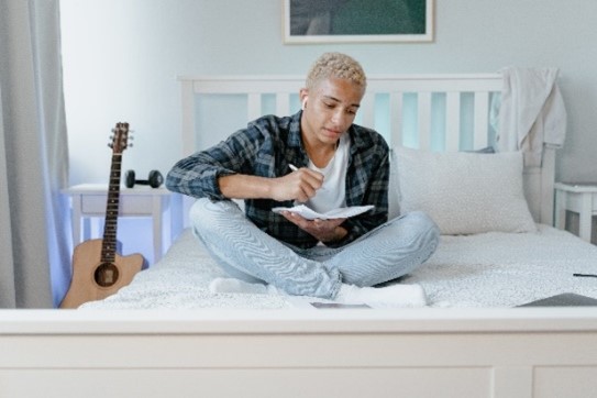 young Black person siting on a bed, writing in a notebook